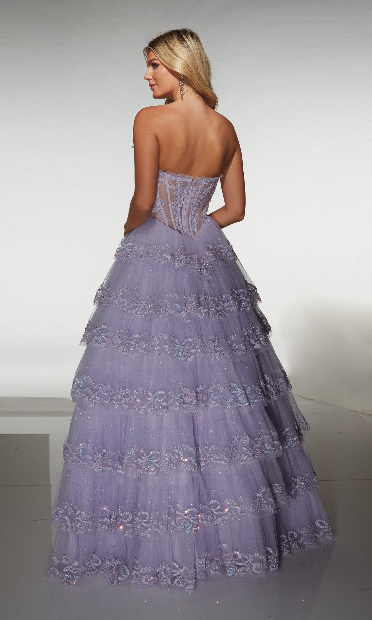 A woman wearing the Alyce 62191 long prom dress, featuring a strapless corset bodice and tiered lavender lace layers, stands facing away from the camera.